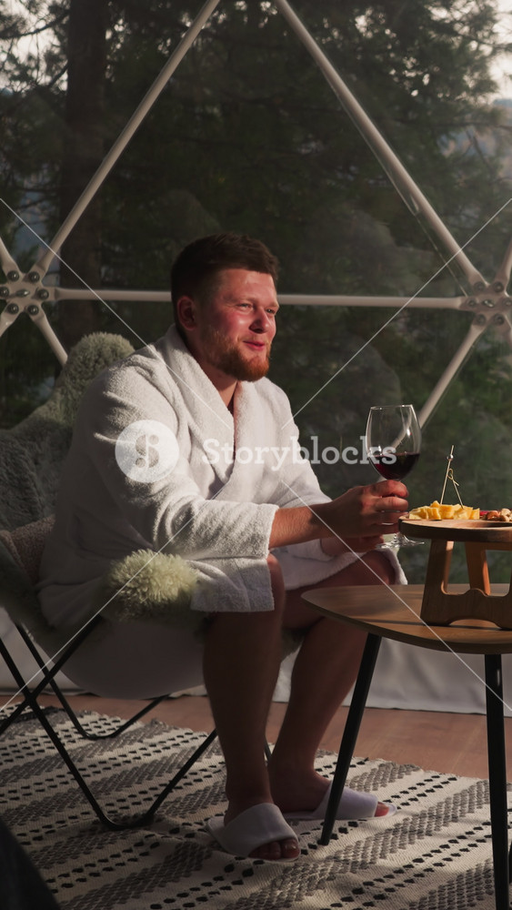 Husband with glass of wine says toast to wife at glamping hotel. Young couple in soft bathrobes celebrates marriage anniversary sitting on