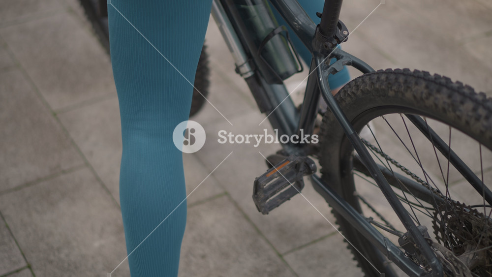 Closeup woman on bicycle pedals, teal leggings and white sneakers push off on city pavement beside dusty rear wheel and chain, quiet morning