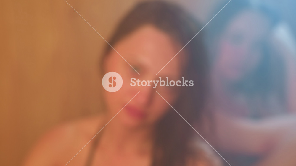 two women wrapped in towels relax inside smoky sauna room, one gazing upward with serene green eyes and soft smile through mist, damp hair