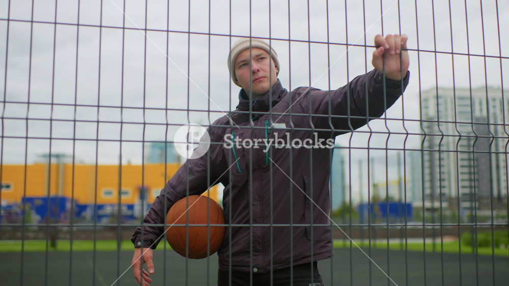 white male leaning on fence holding basketball, moody urban court with yellow warehouse in background and overcast sky. wearing beanie and