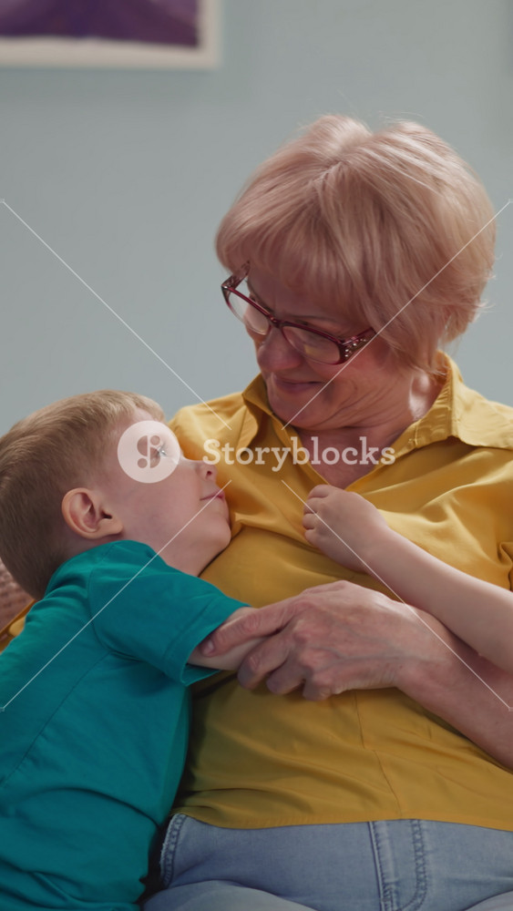 Smiling aged blonde lady cuddles toddler grandson while girl hugs granny in living room. Cute children visit loving grandmother. Domestic