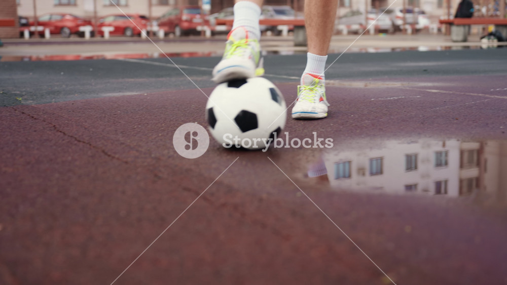 wet urban football feet near puddle, closeup of player controlling ball on rainsoaked asphalt, cleats and sneakers visible, reflection of
