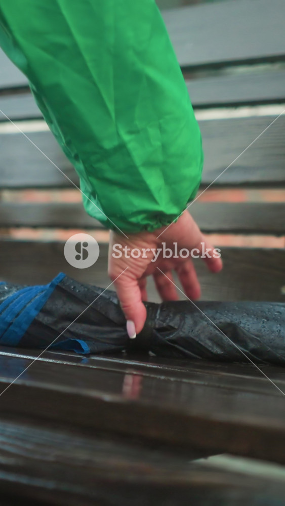 Close-up of individual in green raincoat reaching out for a wet umbrella placed on a wooden swing bench, with a blurred building in the