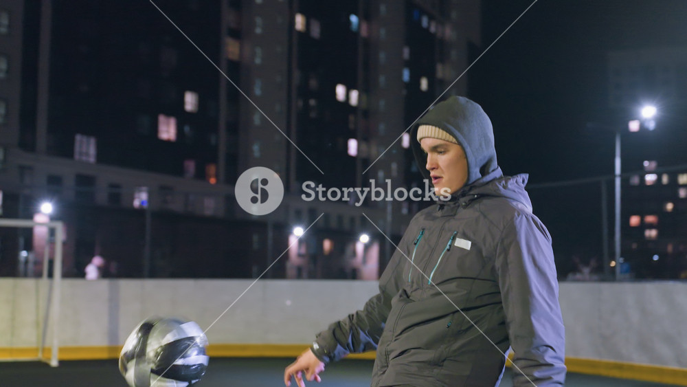 Footballer in hoodie joggling football during nighttime practice on urban sports field surrounded by illuminated high-rise buildings and