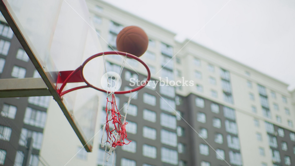 urban basketball hoop city building backdrop, ball arcs toward rim then empties net, red metal rim with frayed chain net, overcast sky