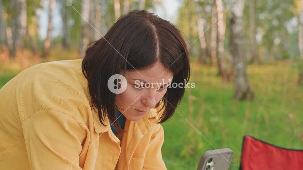 Woman creating social content outdoors. Caucasian woman engaged with her phone in nature setting. Woman in yellow jacket reviews her social