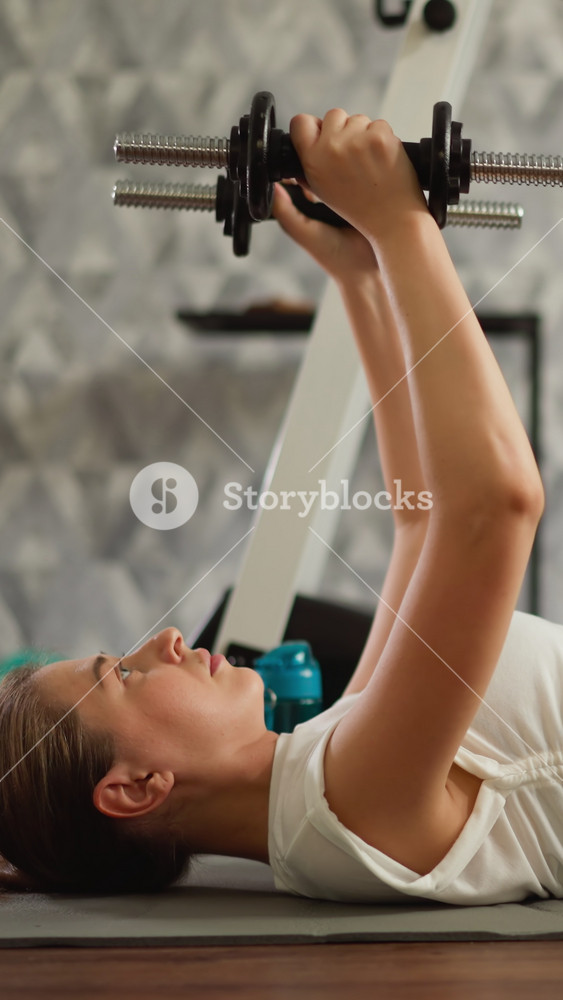 Lady lifts dumbbells extending arms within confines of home. Dedicated young athlete focuses on toning arms with sport equipment in home