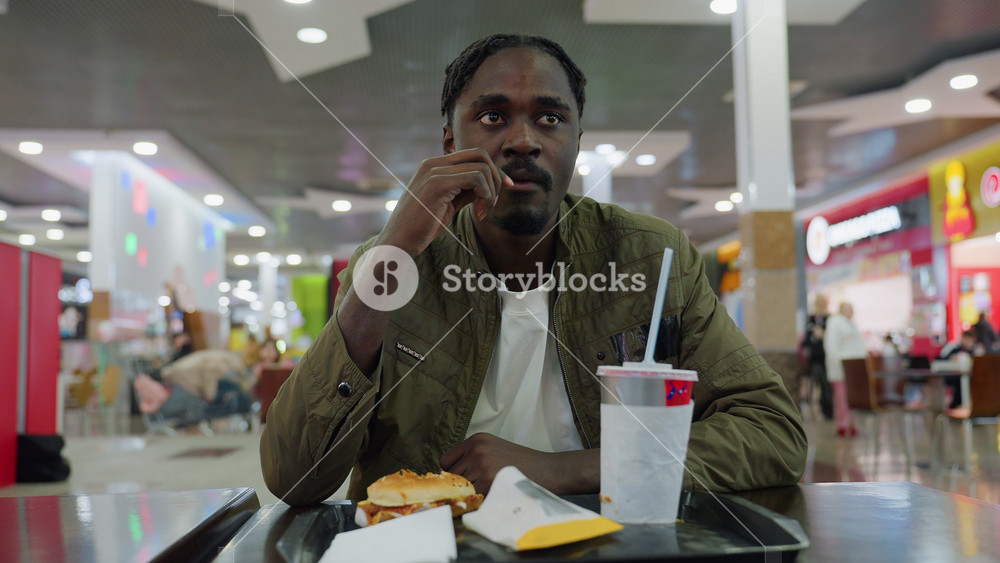 Focused young man wearing green jacket chewing french fries in vibrant food court setting, seated at table with burger, fries, paper cup on