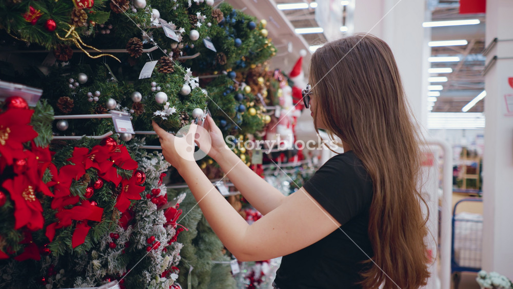 Young woman with and long brown hair in black shirt browsing festive wreaths decorated with ornaments and pinecones in holiday section of