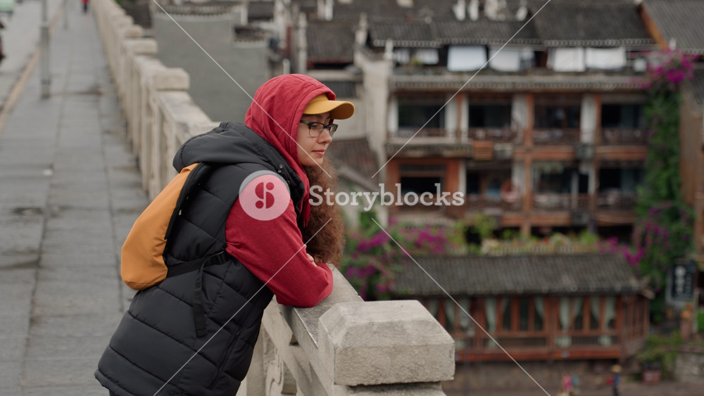 Asian woman leaning on bridge railing overlooking riverside village, wearing red hoodie, black puffer vest and yellow cap with orange