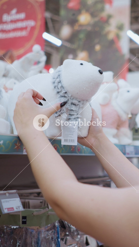 Side view of young woman with long brown hair wearing black shirt and glasses admiring white plush polar bear toy with scarf on store shelf