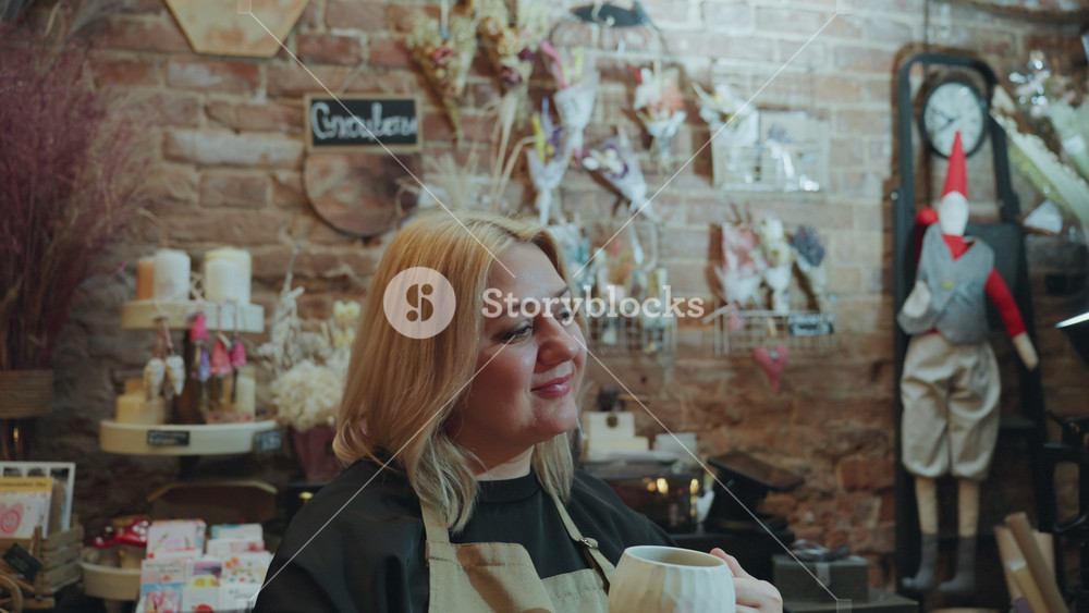 Blonde florist chatting behind shop counter, woman in apron smiling among dried bouquets and quirky decor, brick wall display with gifts and