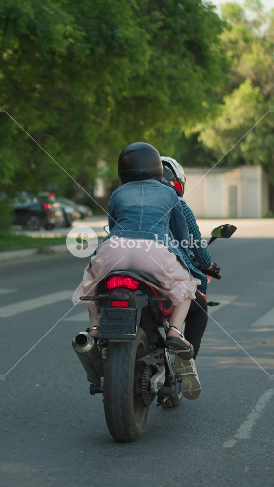 Two women ride a power bike on an urban road. The passenger holds onto the rider as they pass a pedestrian signpost, trees line the road