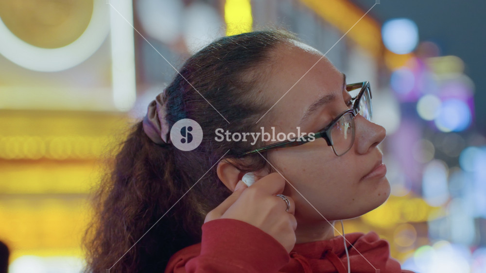 Young woman wearing glasses listening to music, neon lit city street background, pensive student in red hoodie adjusts earbuds, soft bokeh
