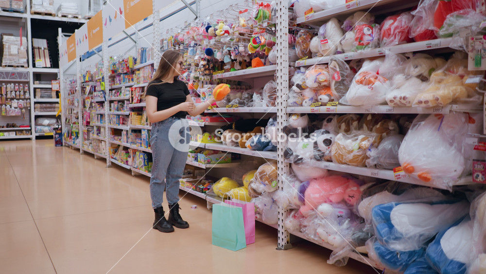 Woman in black top and jeans shopping in toy store, reaching for soft plush toy on shelf, colorful stuffed animals and games line the aisles