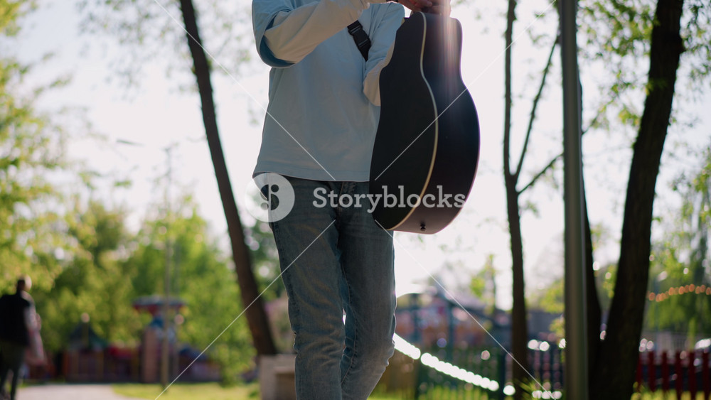 young male guitarist carrying acoustic guitar along treelined path, backlit morning glow filters through leaves wearing casual jeans and
