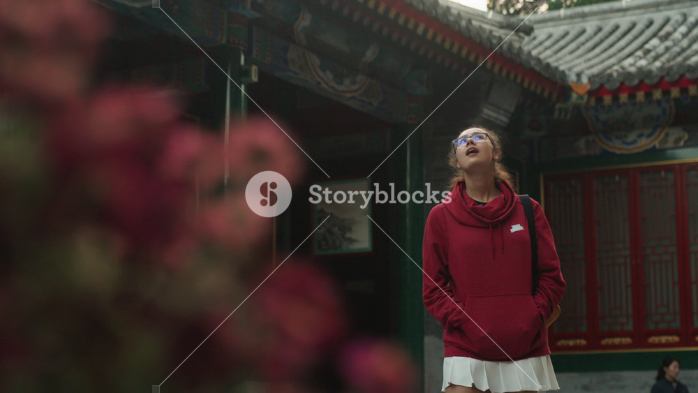 Young woman gazes at temple roof, red hoodie traveler looks upward through blurred blooms, ponytail silhouette against ornate roofline