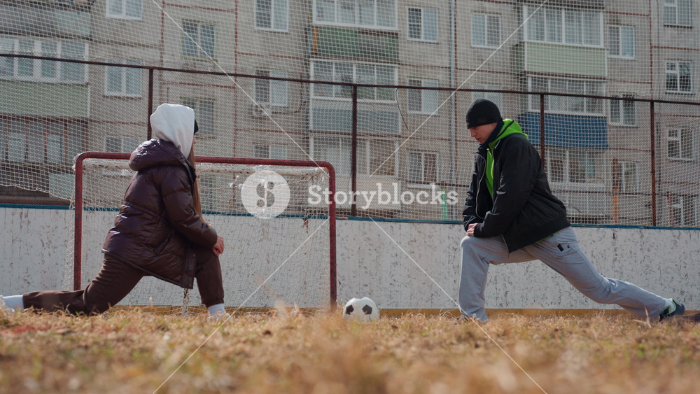 white female challenger stretches near goal while white male opponent readies shot, cold urban pitch, casual winter clothing, playful