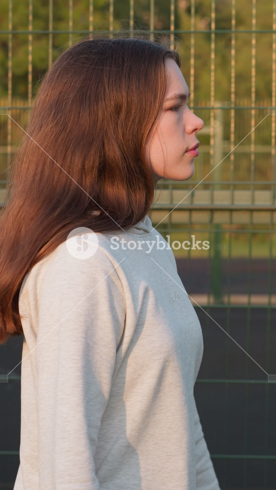 Woman walks by fence of city stadium. Young lady goes to fitness class to improve physical health thinking about problems at sunset. Feeling