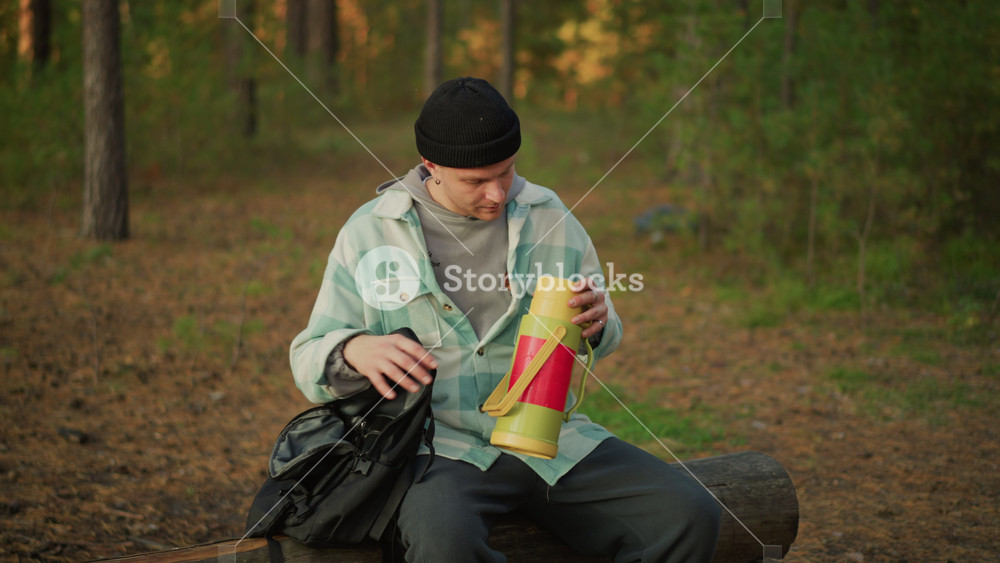 caucasian camper pouring hot drink, yellow thermos and cup on wooden bench, pine woodland setting, steaming beverage, casual plaid shirt and
