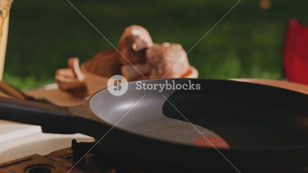 Close up camping pan oil pour hands adding oil to skillet near mushrooms and red camping chair, tranquil morning light, outdoor prep by