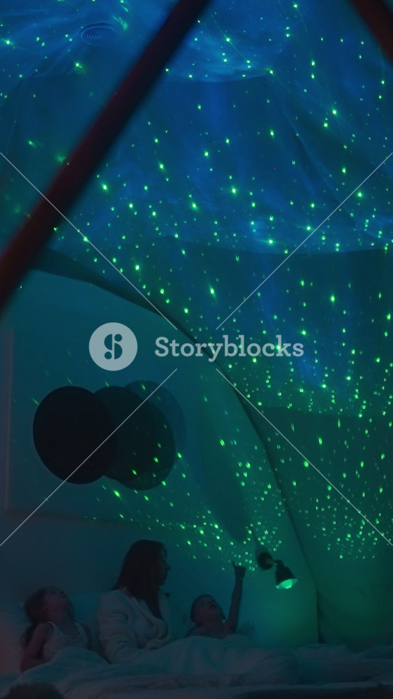 Mother with children lies on white bed and explains locations of stars and planets pointing to projection of sky on white ceiling. Family