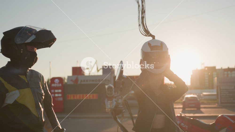 Lady puts on helmet and buckles strap while male rider watches nearby during sunset in front of shopping mall background motorcycles parked