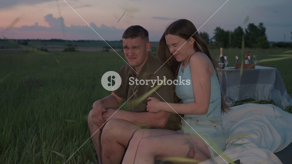 couple sitting on cushioned blanket in open farmland enjoying sunset picnic reacts with laughter and surprise as blades of grass fly around