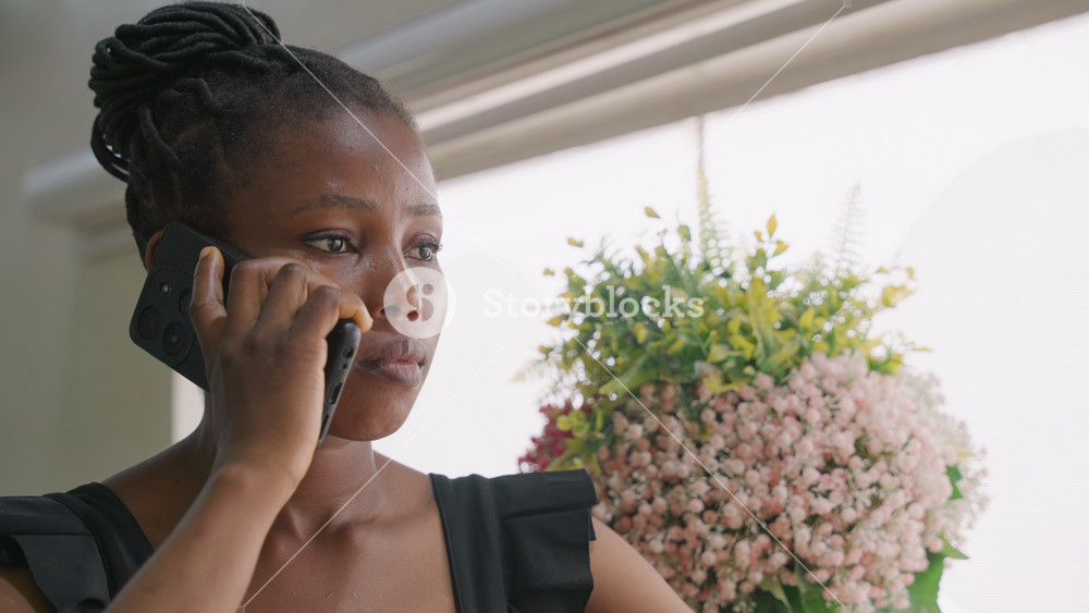 Black woman smiling on phone near pink bouquet, playful flirty mood, bright indoor light, braided hair, planning evening date, laughing