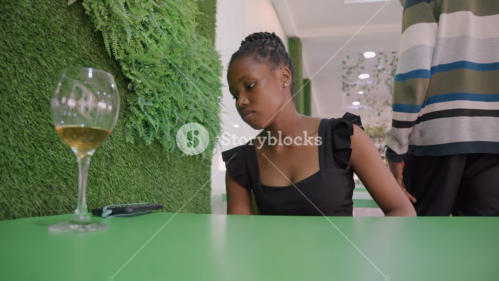 Woman reflective after date with wine glass on table, green living wall and empty chair create quiet backdrop, female in black dress watches