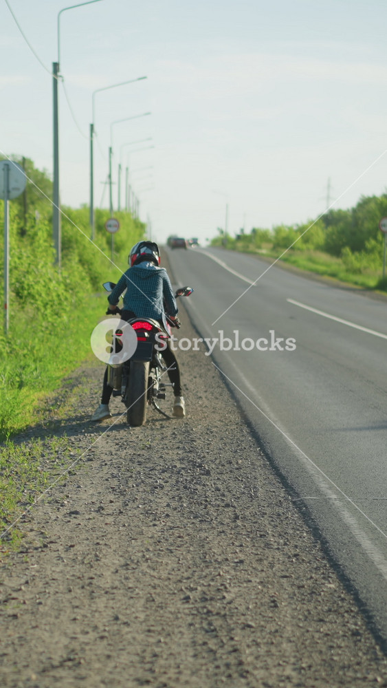 A back view of a woman climbing onto a motorcycle parked by the roadside, with lush green bushes in the background and cars approaching from