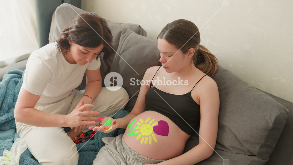Pregnant caucasian woman belly painting with supportive doula friend, colorful flower and heart body art on exposed bump, cozy living room