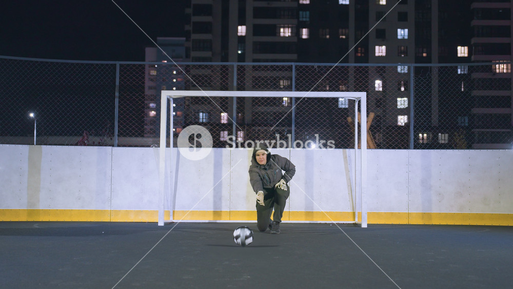 Goalkeeper in hoodie rolling soccer ball on ground during nighttime practice on outdoor field, fenced urban sports area with city lights in