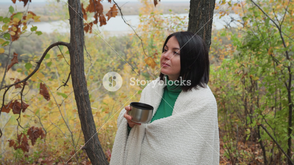 white woman sipping from mug in woodland wrapped in blanket, green turtleneck and warm cup, autumn foliage and quiet river suggestion