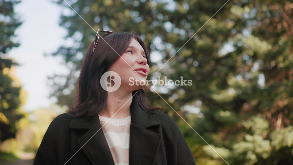 fashionable girl under autumn canopy gazes upward with warm smile, admiring nature in sunny park, wearing dark coat and striped sweater