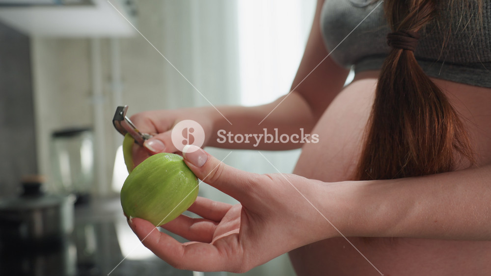 Midshot pregnant woman holding green fruit near belly at countertop, examining skin and stem, mindful nutrition choice, soft morning light