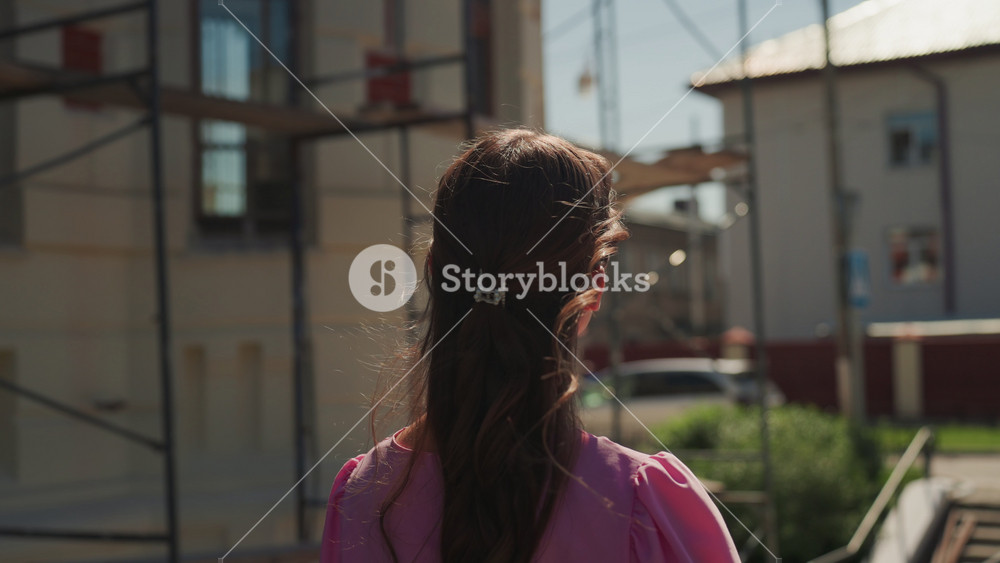 Woman from behind on street, sunlit backview with glittering droplets in hair casual pink dress, city traffic in background, passerby mood