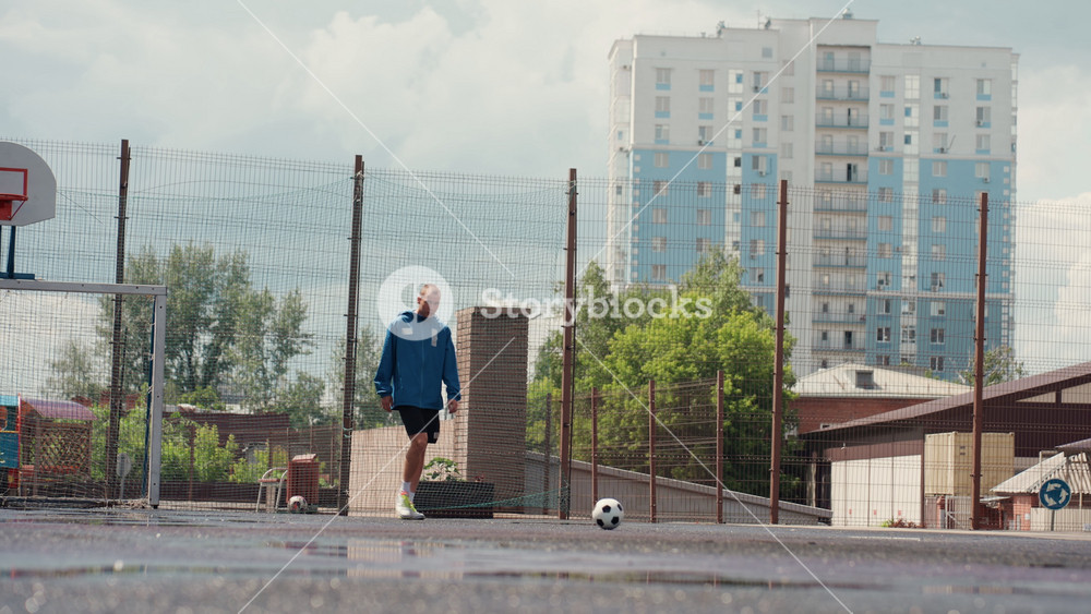 street footballer practicing alone on wet court, overcast sky, urban highrise backdrop, fence topped with barbed wire, puddles splashing as