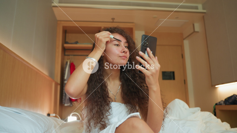 Young woman hotel bed applying makeup while checking phone mirror, casual travel scene showing curly hair and white towel dress, natural