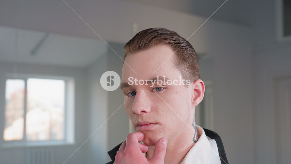 portrait view of stylish man with slicked hair and neck tattoo holding chin in thoughtful pose, standing calmly near large wall mirror