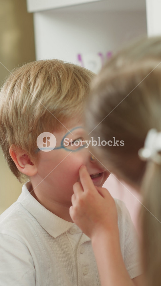 Boy laughs while girl paints face slow motion. Sister draws glasses and lines around nose. Friendly couple of kids plays with body painting