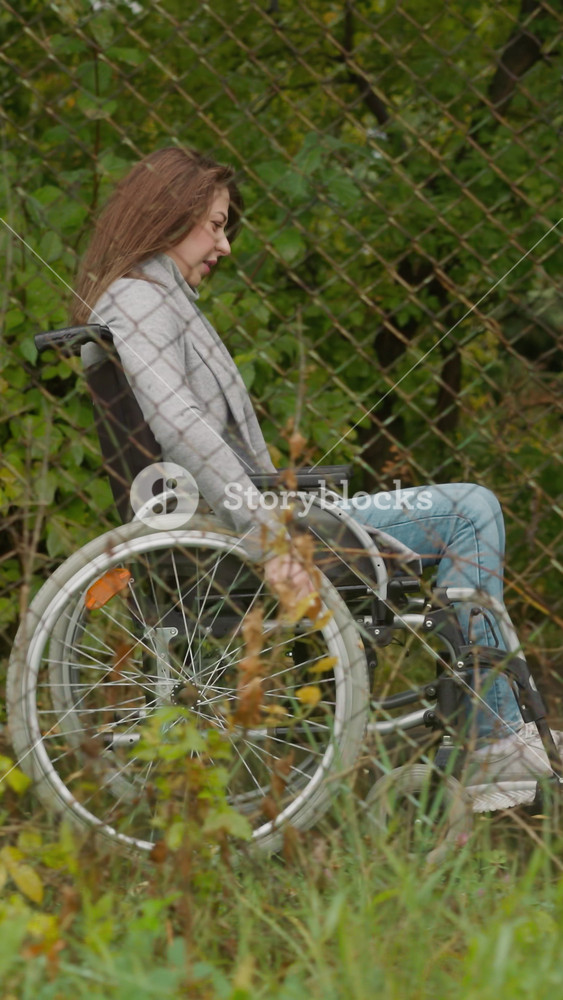 Young woman in wheelchair rests moving in park past old mesh fence. Health restoration at resort complex. Rehabilitation after legs injury
