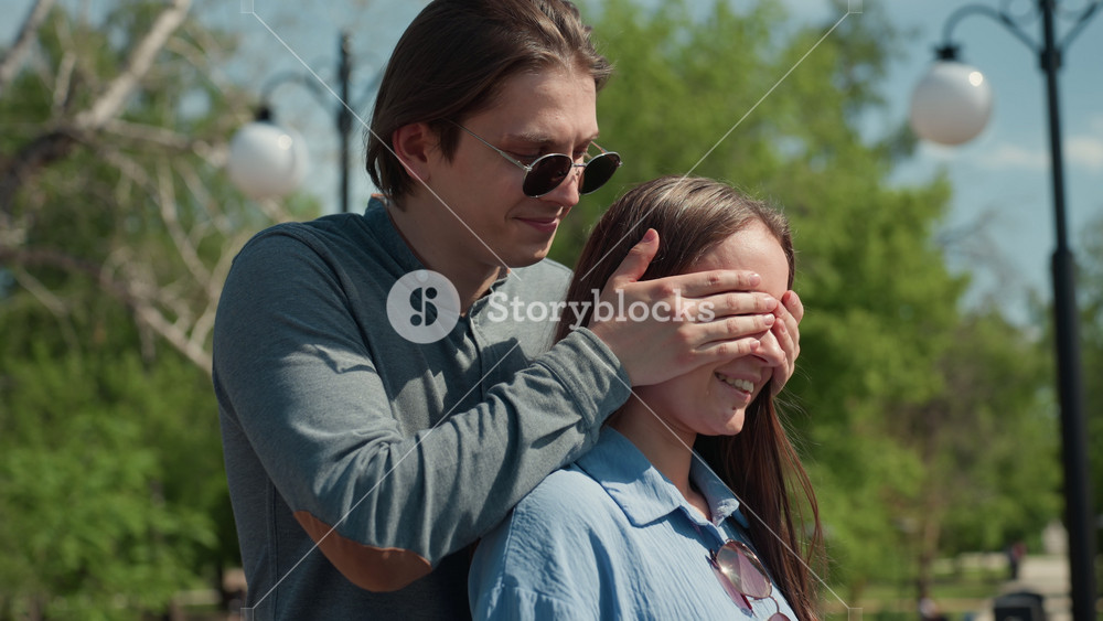 sunglasses man surprises white girlfriend outdoors with tender cover of eyes and gentle kiss, public park scene with lamp posts and walking