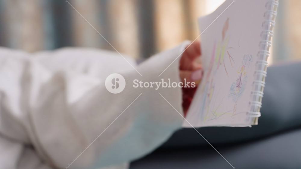 Hands holding sketchbook with colorful child drawing of house, tree, sun and people, finger pointing at detail of artwork, cozy home