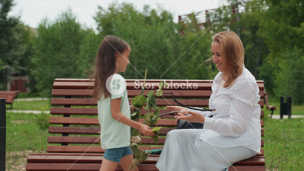 Mother and daughter sharing bench gift moment, child offers leafy branch while parent smiles and reaches, warm family connection, casual