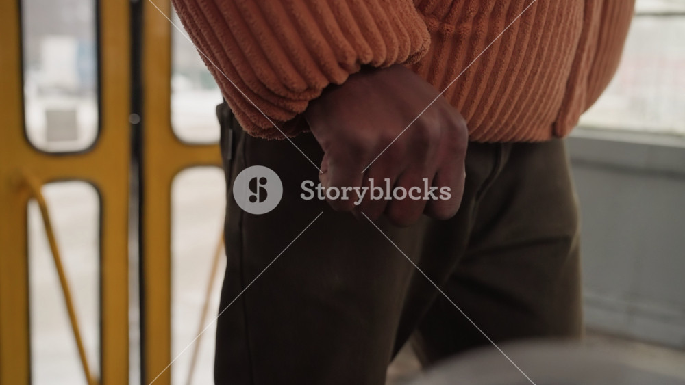 Black commuter standing on bus, orange knit sweater and brown pants, close detail of hand gripping metal pole by yellow door, urban interior