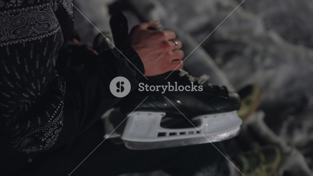 Close up of person gripping black hockey skate boot by hand, detailed view of blade and lace against blurred outdoor ice rink surface, cold