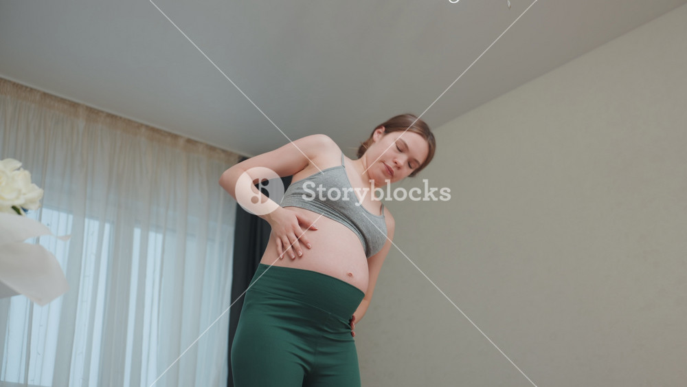 Pregnant caucasian woman in living room gently stretching and easing lower back pain while wearing grey sports bra and green leggings, soft