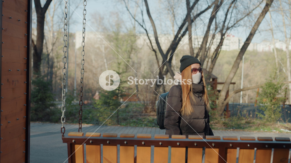 Park railing caucasian woman in mask standing behind wooden barrier, long red hair under beanie, sunglasses and winter coat, watchful