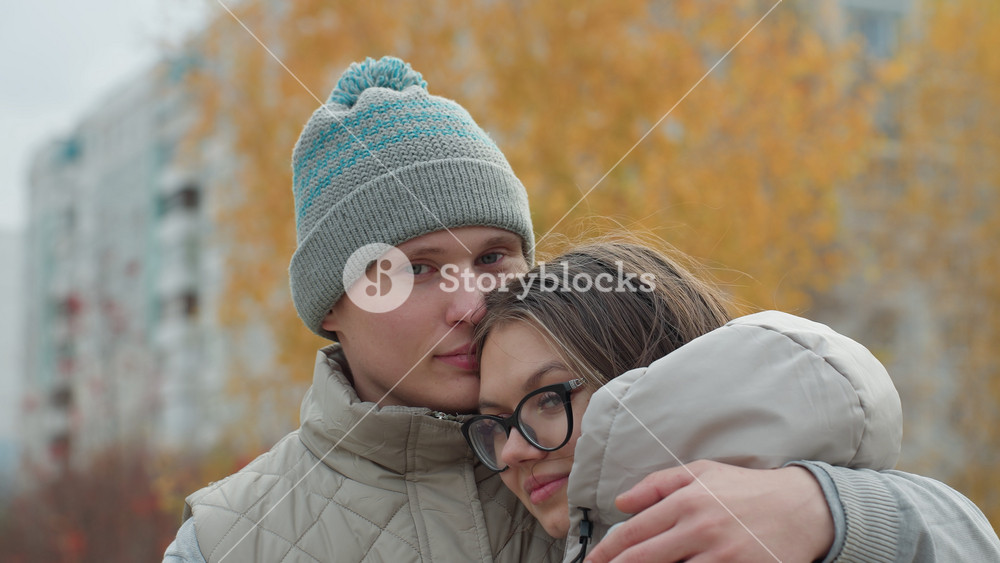 Close up of two friends embracing warmly in identical winter outfits outdoors with soft view of golden autumn trees and buildings in